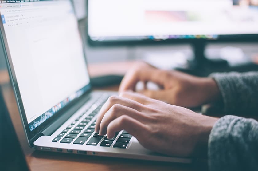 A person working on a laptop, with a second screen in the background.